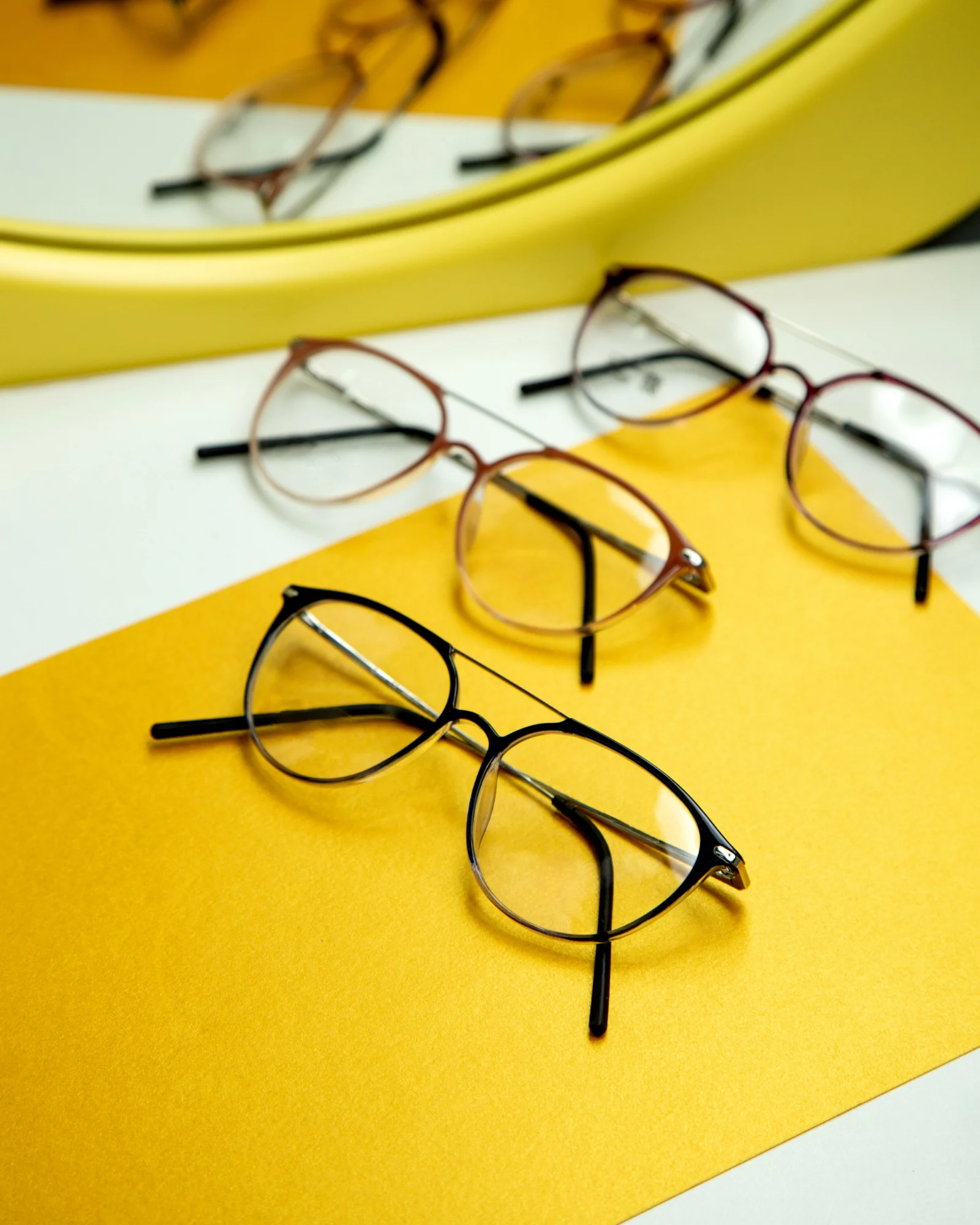 Montures de lunettes tendance posées sur une table colorée Trois paires de lunettes de vue modernes posées sur une surface jaune avec un miroir en arrière-plan